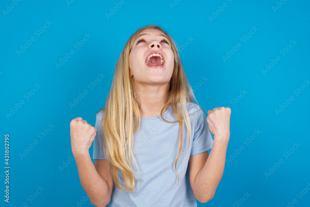 beautiful Caucasian little girl wearing blue T-shirt over blue background looks with excitement up, keeps hands raised, notices something unexpected. beautiful Caucasian little girl wearing blue T-shirt over blue background looks with excitement up, keeps hands raised, notices something unexpected.