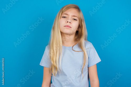 Gloomy, bored beautiful Caucasian little girl wearing blue T-shirt over blue background frowns face looking up, being upset with so much talking hands down, feels tired and wants to leave. © Jihan
