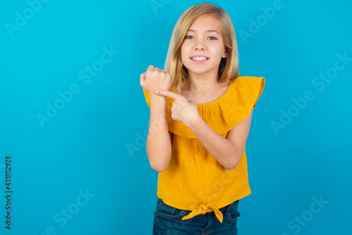 Caucasian kid girl wearing yellow T-shirt against blue wall In hurry pointing to wrist watch, impatience, looking at the camera with relaxed expression