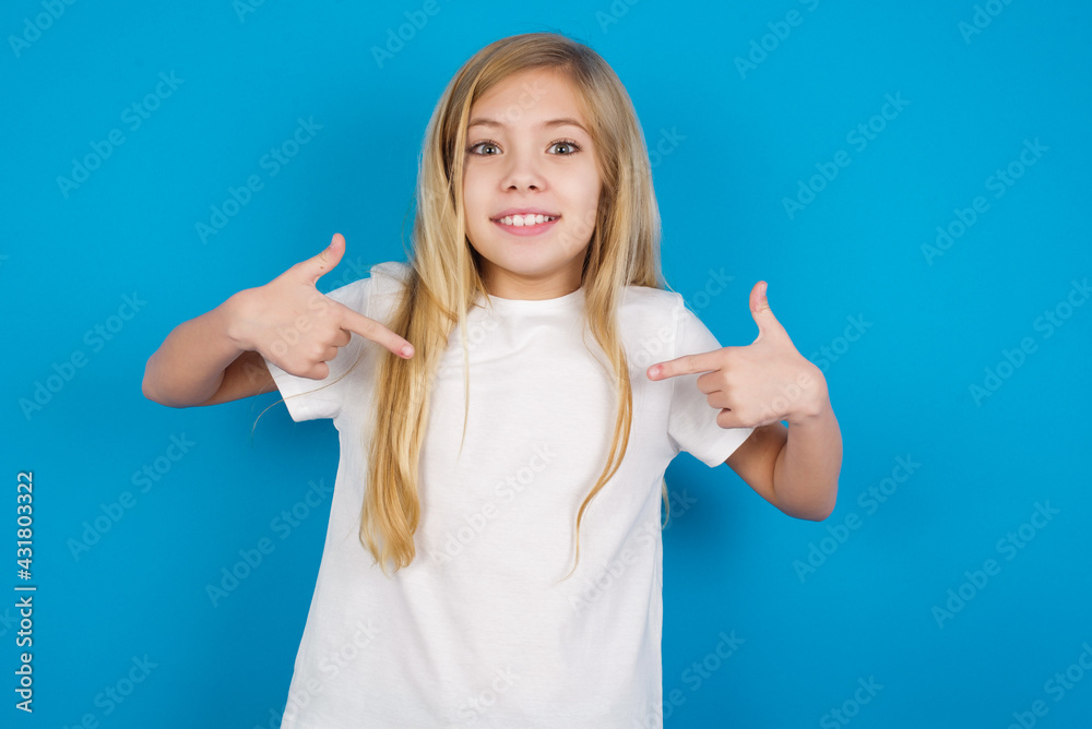 Pick me! Confident, self-assured and charismatic beautiful Caucasian little girl wearing white T-shirt over blue background promoting oneself as wanting role smiling broadly and pointing at body. Pick me! Confident, self-assured and charismatic beautiful Caucasian little girl wearing white T-shirt over blue background promoting oneself as wanting role smiling broadly and pointing at body.