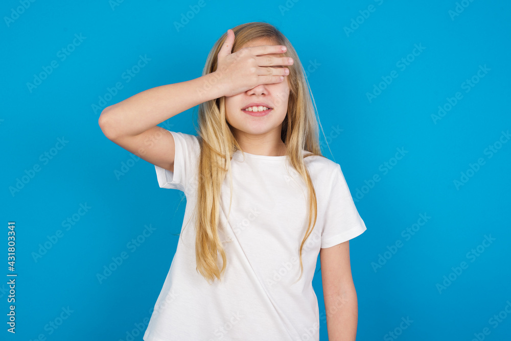 Happy beautiful Caucasian little girl wearing white T-shirt over blue wall closing eyes with hand going to see surprise prepared by friend standing and smiling in anticipation for something wonderful. Happy beautiful Caucasian little girl wearing white T-shirt over blue wall closing eyes with hand going to see surprise prepared by friend standing and smiling in anticipation for something wonderful.