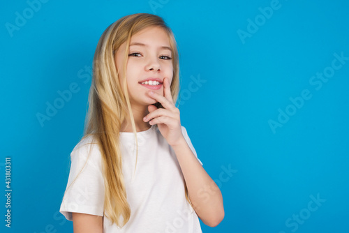 Carefree successful beautiful Caucasian little girl wearing white T-shirt over blue background touching jawline gazing camera tilting head grinning white teeth delighted. Dental care concept.