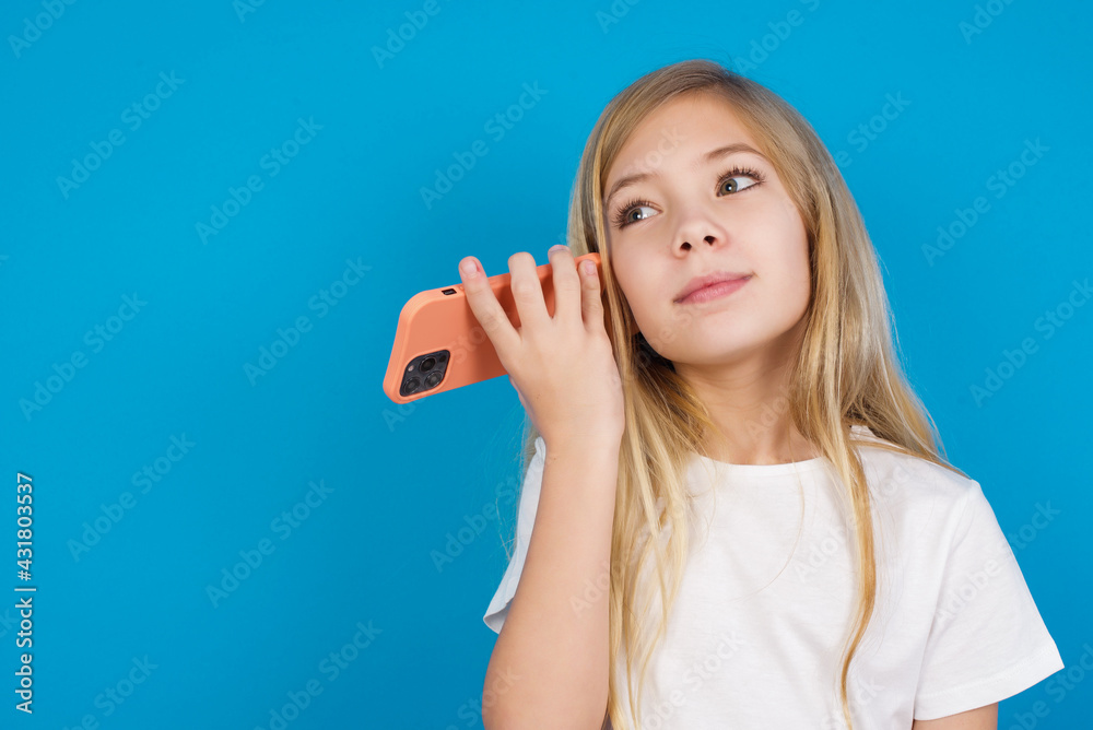 Smiling beautiful Caucasian little girl wearing white T-shirt over blue background listening a voice message from her smartphone. Communication and technology concept. Smiling beautiful Caucasian little girl wearing white T-shirt over blue background listening a voice message from her smartphone. Communication and technology concept.