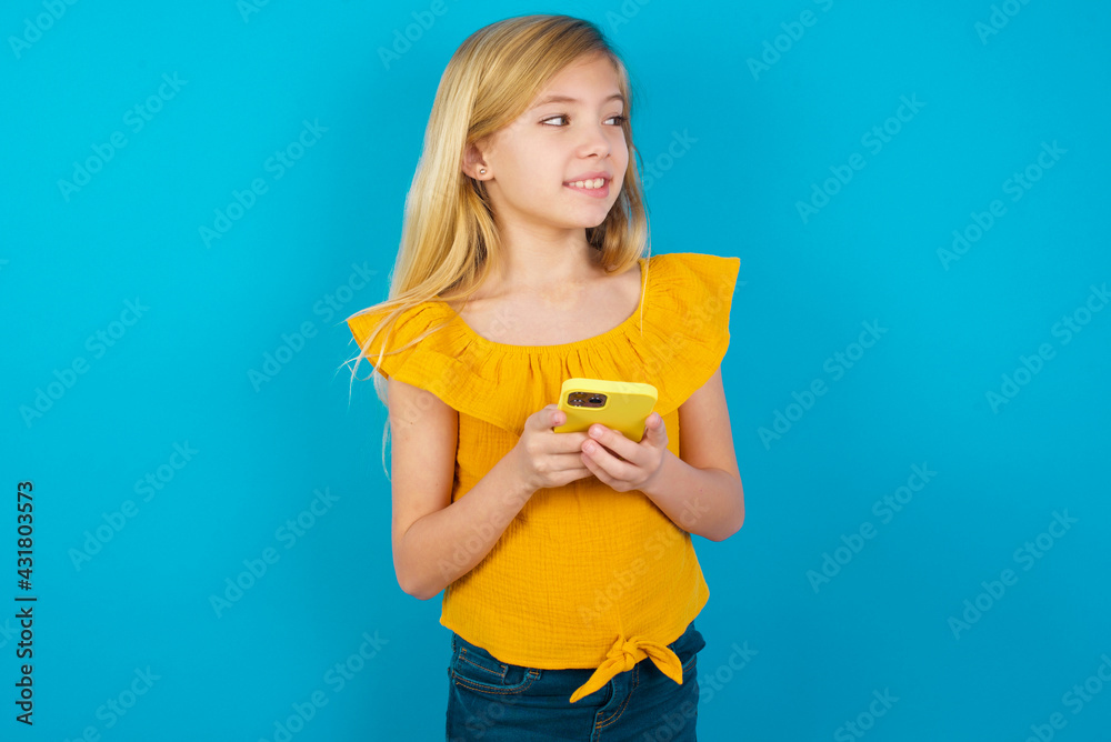Caucasian kid girl wearing yellow T-shirt against blue wall hold telephone hands read good youth news look empty space advert Caucasian kid girl wearing yellow T-shirt against blue wall hold telephone hands read good youth news look empty space advert