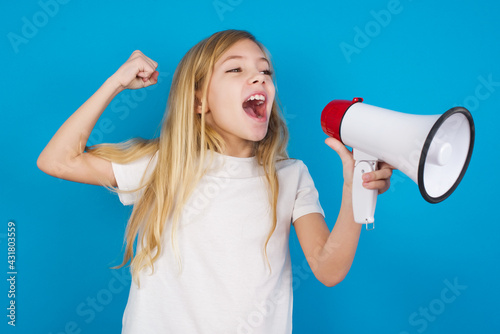 beautiful Caucasian little girl wearing white T-shirt over blue background communicates shouting loud holding a megaphone, expressing success and positive concept, idea for marketing or sales.