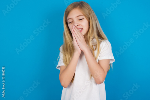 Overjoyed beautiful Caucasian little girl wearing white T-shirt over blue background laughs joyfully and keeps palms pressed together hears something funny