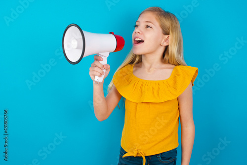 Caucasian kid girl wearing yellow T-shirt against blue wall Through Megaphone with Available Copy Space