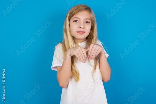 beautiful Caucasian little girl wearing white T-shirt over blue background makes bunny paws and looks with innocent expression plays with her little kid
