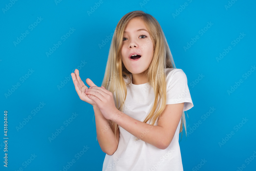 Fototapeta premium Surprised emotional beautiful Caucasian little girl wearing white T-shirt over blue background rubs palms and stares at camera with disbelief