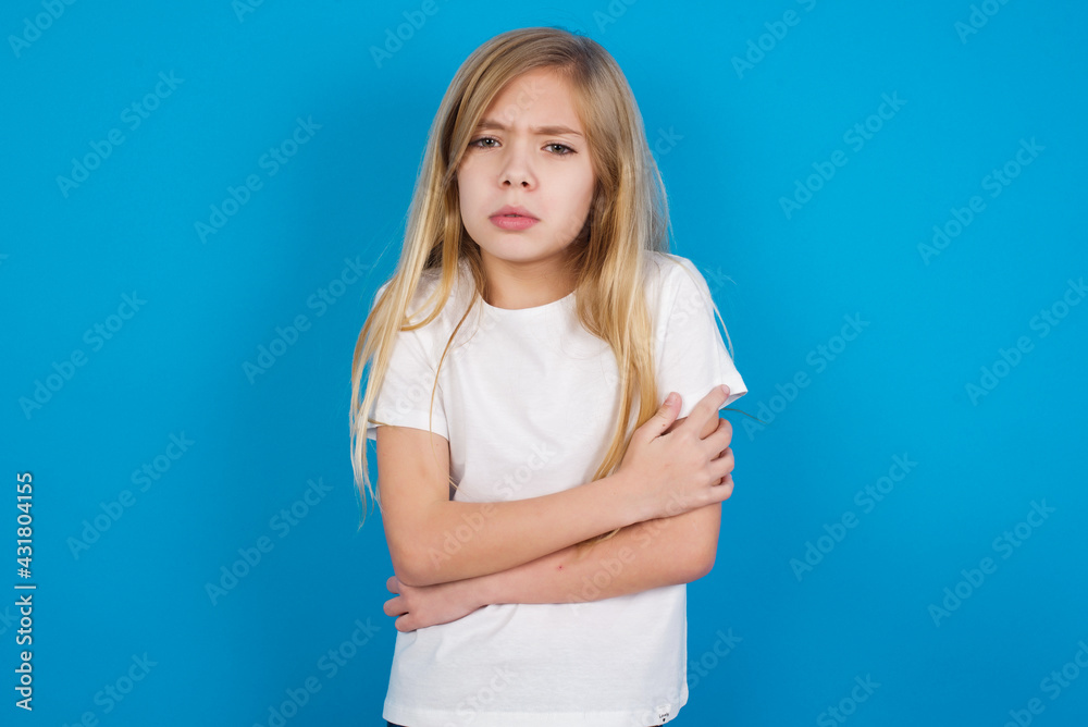 beautiful Caucasian little girl wearing white T-shirt over blue background shaking and freezing for winter cold with sad and shock expression on face. beautiful Caucasian little girl wearing white T-shirt over blue background shaking and freezing for winter cold with sad and shock expression on face.