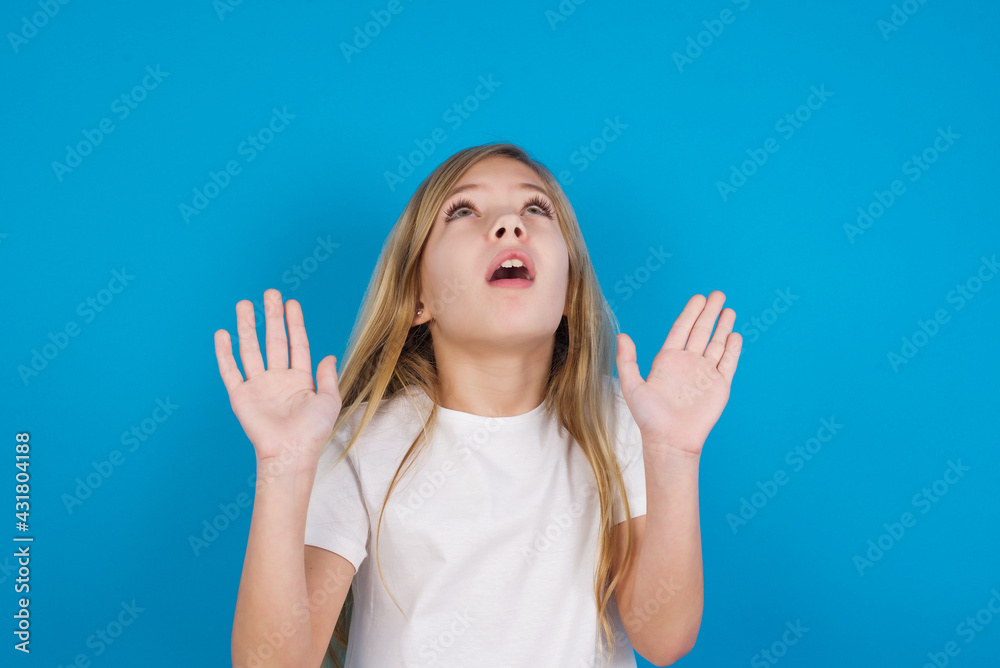 beautiful Caucasian little girl wearing white T-shirt over blue background keeps palms forward and looks with fright above on ceiling tries to defense herself from invisible danger opens mouth. beautiful Caucasian little girl wearing white T-shirt over blue background keeps palms forward and looks with fright above on ceiling tries to defense herself from invisible danger opens mouth.