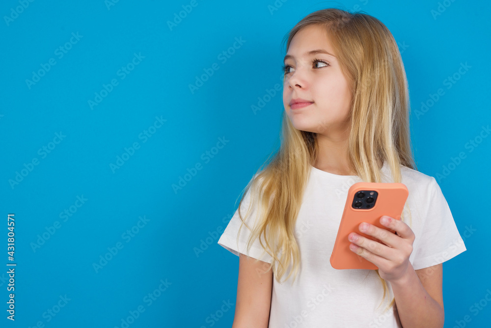 beautiful Caucasian little girl wearing white T-shirt over blue background holds mobile phone uses high speed internet and social networks has online communication. Modern technologies concept beautiful Caucasian little girl wearing white T-shirt over blue background holds mobile phone uses high speed internet and social networks has online communication. Modern technologies concept