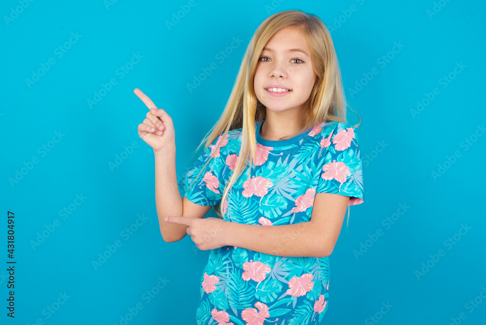 Caucasian kid girl wearing hawaiian T-shirt against blue wall points at copy space indicates for advertising gives right direction Caucasian kid girl wearing hawaiian T-shirt against blue wall points at copy space indicates for advertising gives right direction