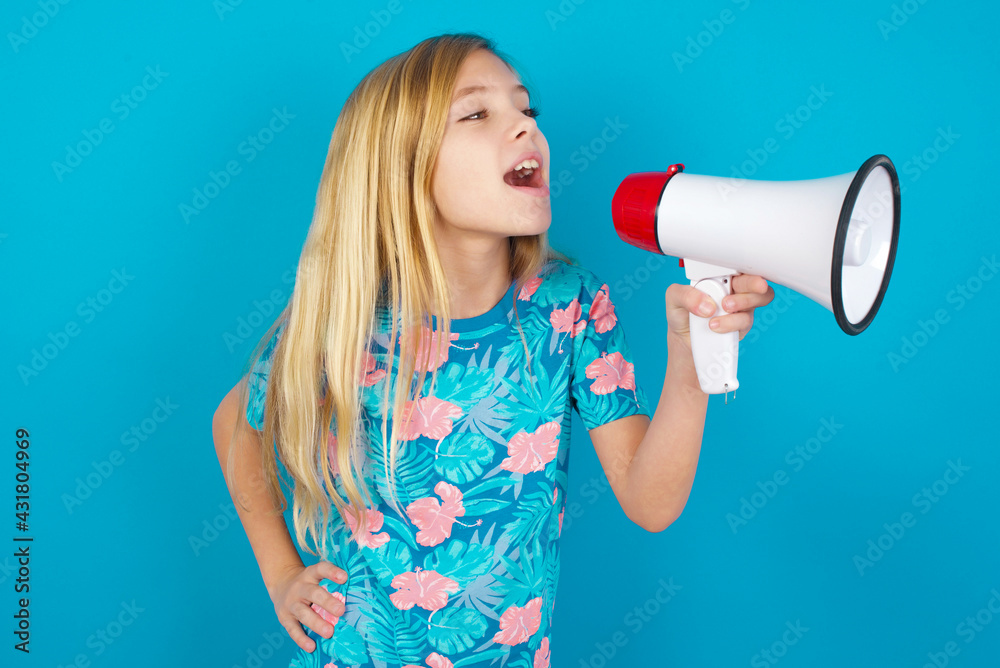Funny Caucasian kid girl wearing hawaiian T-shirt against blue wall People sincere emotions lifestyle concept. Mock up copy space. Screaming in megaphone. Funny Caucasian kid girl wearing hawaiian T-shirt against blue wall People sincere emotions lifestyle concept. Mock up copy space. Screaming in megaphone.