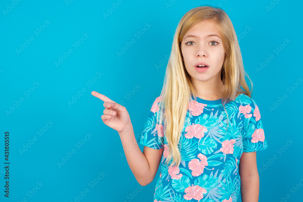 Emotive Caucasian kid girl wearing hawaiian T-shirt against blue wall keeps jaw dropped from shock demonstrates amazing promo points right on blank space demonstrates big shopping sale. Advertisement Emotive Caucasian kid girl wearing hawaiian T-shirt against blue wall keeps jaw dropped from shock demonstrates amazing promo points right on blank space demonstrates big shopping sale. Advertisement