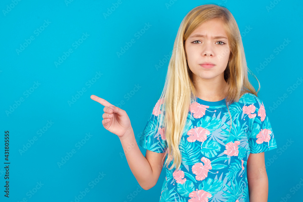 Serious Caucasian kid girl wearing hawaiian T-shirt against blue wall smirks face points away on copy space shows something unpleasant. Look at this advertisement. Big price concept Serious Caucasian kid girl wearing hawaiian T-shirt against blue wall smirks face points away on copy space shows something unpleasant. Look at this advertisement. Big price concept