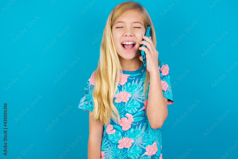 Overemotive happy Caucasian kid girl wearing hawaiian T-shirt against blue wall laughs out positively hears funny story from friend during telephone conversation Overemotive happy Caucasian kid girl wearing hawaiian T-shirt against blue wall laughs out positively hears funny story from friend during telephone conversation