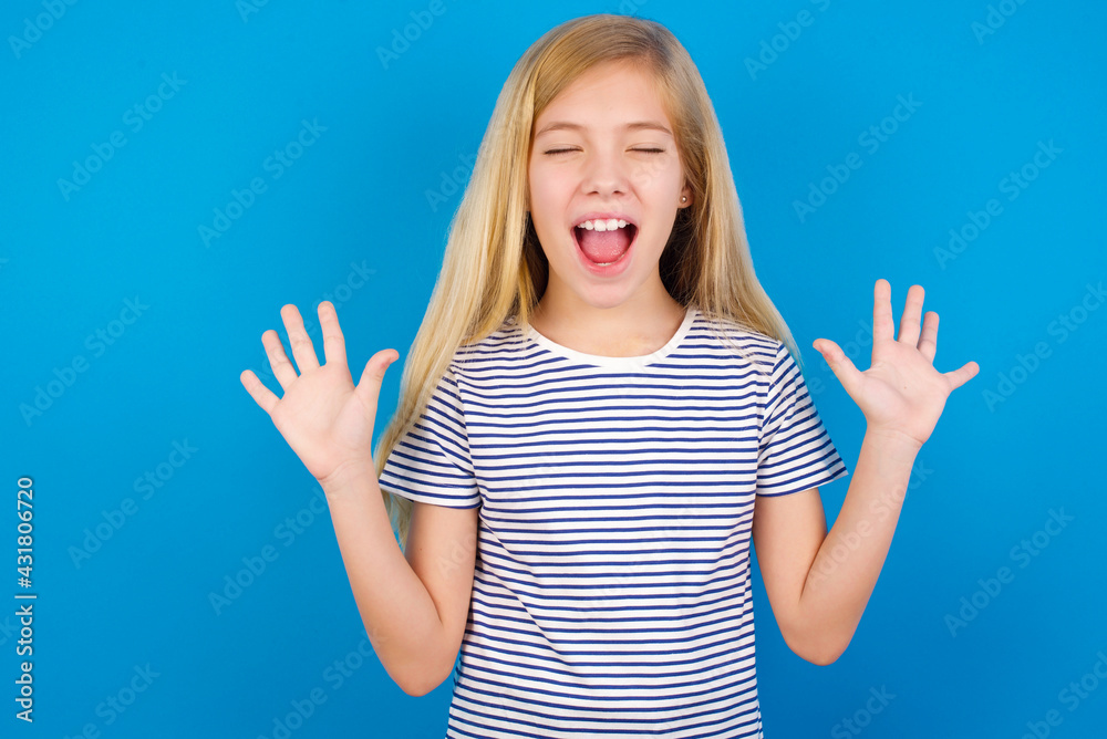 Fototapeta premium Emotive Caucasian kid girl wearing striped shirt against blue wall laughs loudly, hears funny joke or story, raises palms with satisfaction, being overjoyed amused by friend