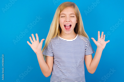 Emotive Caucasian kid girl wearing striped shirt ​against blue wall laughs loudly, hears funny joke or story, raises palms with satisfaction, being overjoyed amused by friend © Roquillo