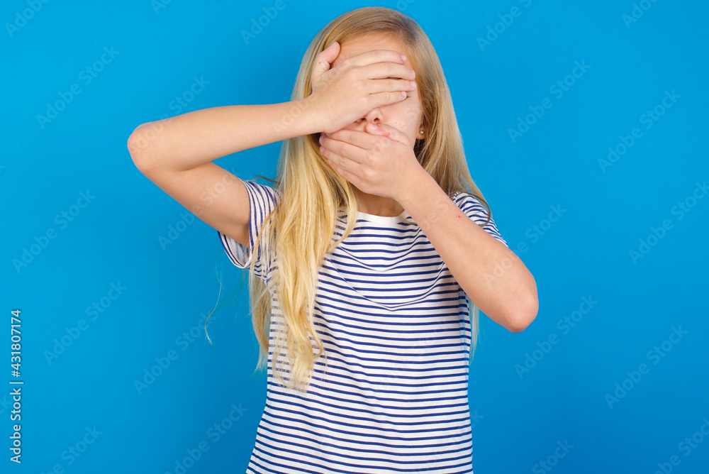 Caucasian kid girl wearing striped shirt against blue wall Covering eyes and mouth with hands, surprised and shocked. Hiding emotions. Caucasian kid girl wearing striped shirt against blue wall Covering eyes and mouth with hands, surprised and shocked. Hiding emotions.