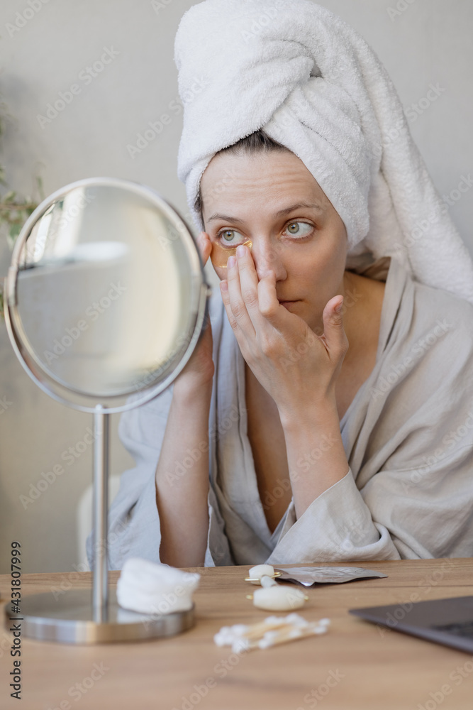 natural beauty and self-care, Caucasian woman with a towel on her hair in front of the mirror applies moisturizing cream or patches on her face, woman takes care of her skin massage face quartz natural beauty and self-care, Caucasian woman with a towel on her hair in front of the mirror applies moisturizing cream or patches on her face, woman takes care of her skin massage face quartz