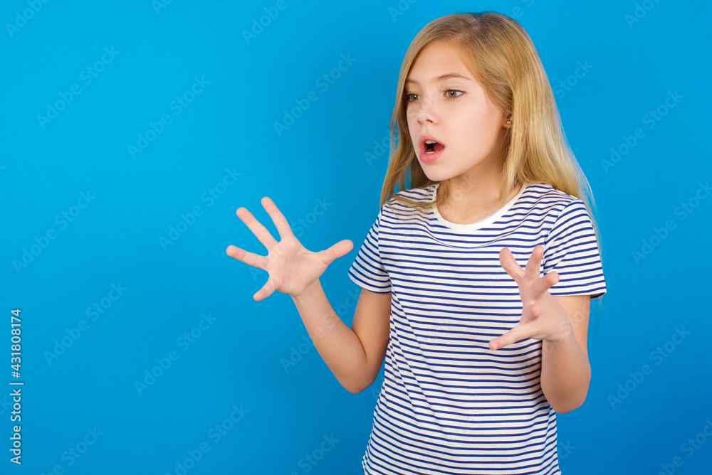Fototapeta premium Caucasian kid girl wearing striped shirt ​against blue wall shouts loud, keeps eyes opened and hands tense.