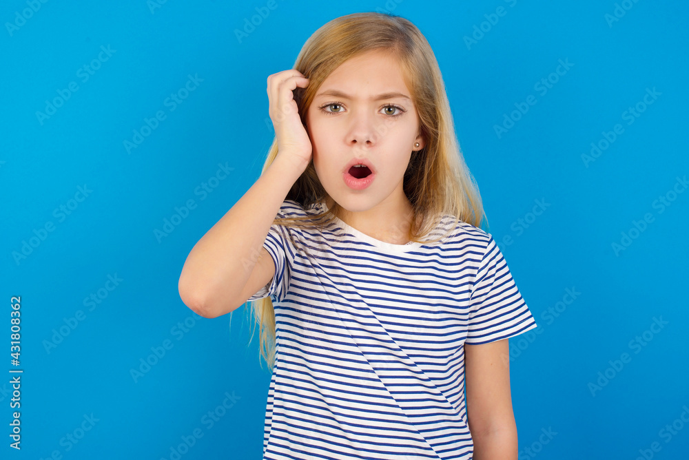 Embarrassed Caucasian kid girl wearing striped shirt against blue wall with shocked expression, expresses great amazement, Puzzled model poses indoor Embarrassed Caucasian kid girl wearing striped shirt against blue wall with shocked expression, expresses great amazement, Puzzled model poses indoor