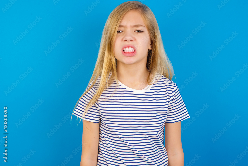 Caucasian kid girl wearing striped shirt against blue wall keeps teeth clenched, frowns face in dissatisfaction, irritated because of much duties. Caucasian kid girl wearing striped shirt against blue wall keeps teeth clenched, frowns face in dissatisfaction, irritated because of much duties.