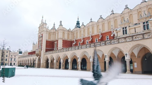 Panoramic footage of Sukiennice Museum, a huge market hall in the middle of Main Square in Old Town Krakow. A flock of pigeons flying. People standing outside the museum. Snowy landscape.  photo