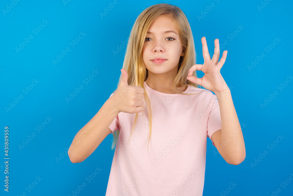 Caucasian kid girl wearing pink shirt against blue wall smiling and looking happy, carefree and positive, gesturing victory or peace with one hand Caucasian kid girl wearing pink shirt against blue wall smiling and looking happy, carefree and positive, gesturing victory or peace with one hand