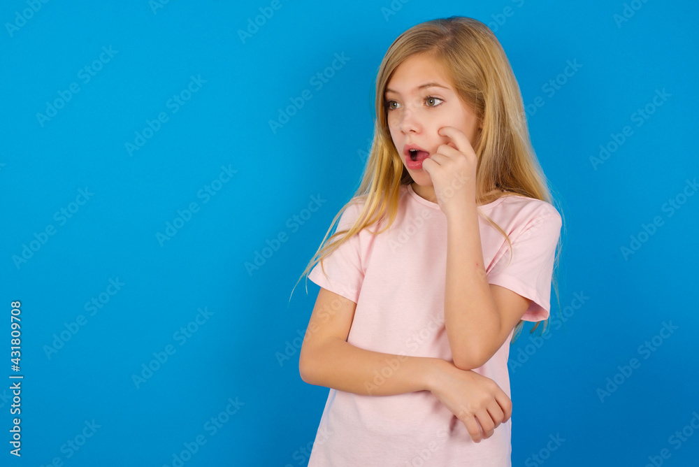 Astonished Caucasian kid girl wearing pink shirt against blue wall looks aside surprisingly with opened mouth. Astonished Caucasian kid girl wearing pink shirt against blue wall looks aside surprisingly with opened mouth.