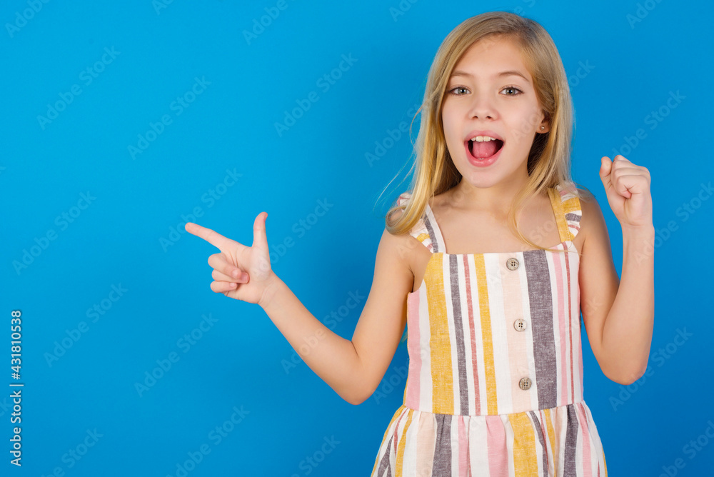 Obraz premium Caucasian kid girl wearing dress against blue wall points at empty space holding fist up, winner gesture.