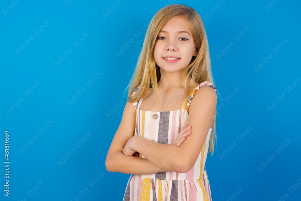 Portrait of Caucasian kid girl wearing dress against blue wall standing with folded arms and smiling Portrait of Caucasian kid girl wearing dress against blue wall standing with folded arms and smiling