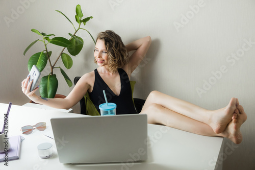Young pretty woman holding fresh summer cocktail and looking away with smile while sitting at workspace, dreaming about vacation. Coronavirus situation in tourism industry. Quarantine. Isolation.
