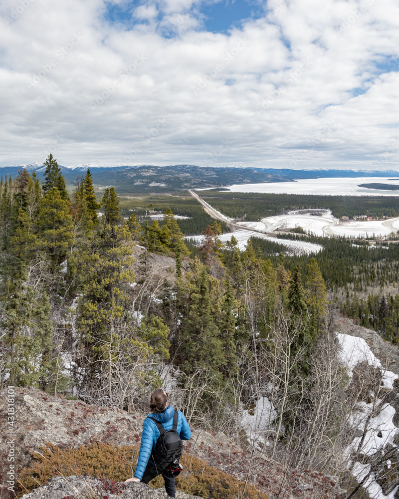Man standing on a rocky surface in northern Canada while gazing into the distance of a stunning scenic view with winding river in the background. Wearing blue jacket, black pants with hiking backpack.