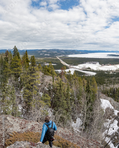 Man standing on a rocky surface in northern Canada while gazing into the distance of a stunning scenic view with winding river in the background. Wearing blue jacket, black pants with hiking backpack.