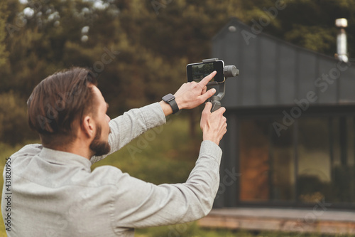Fototapeta Back view of millennial man recording video at smartphone with tripod
