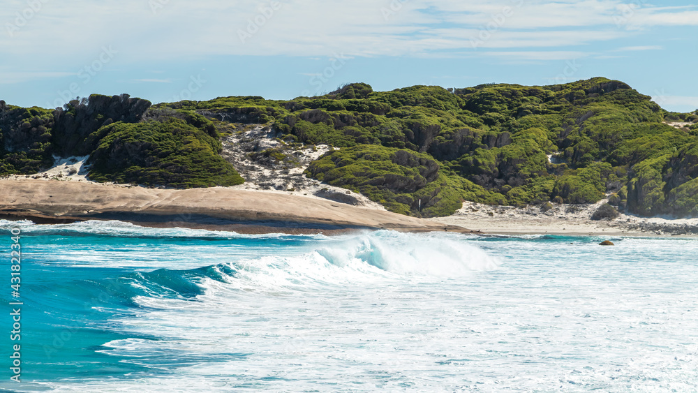 Landscape view of waves breaking over smooth granie rocks at Blue Haven Beach near Esperance in Western Australia under a bright clear blue sky.