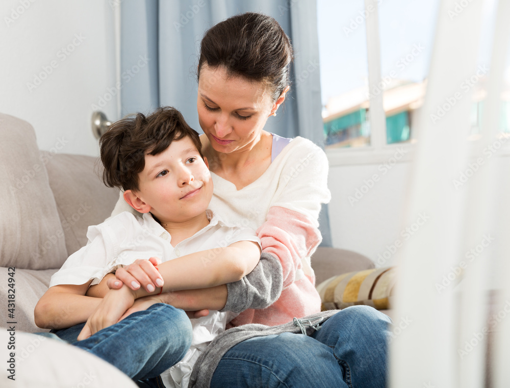 Cheerful teen boy with his mother embracing while sitting on sofa at home..