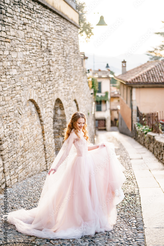 Smiling bride in a beautiful long pink dress stands on the cobblestones of an old street in Bergamo, Italy
