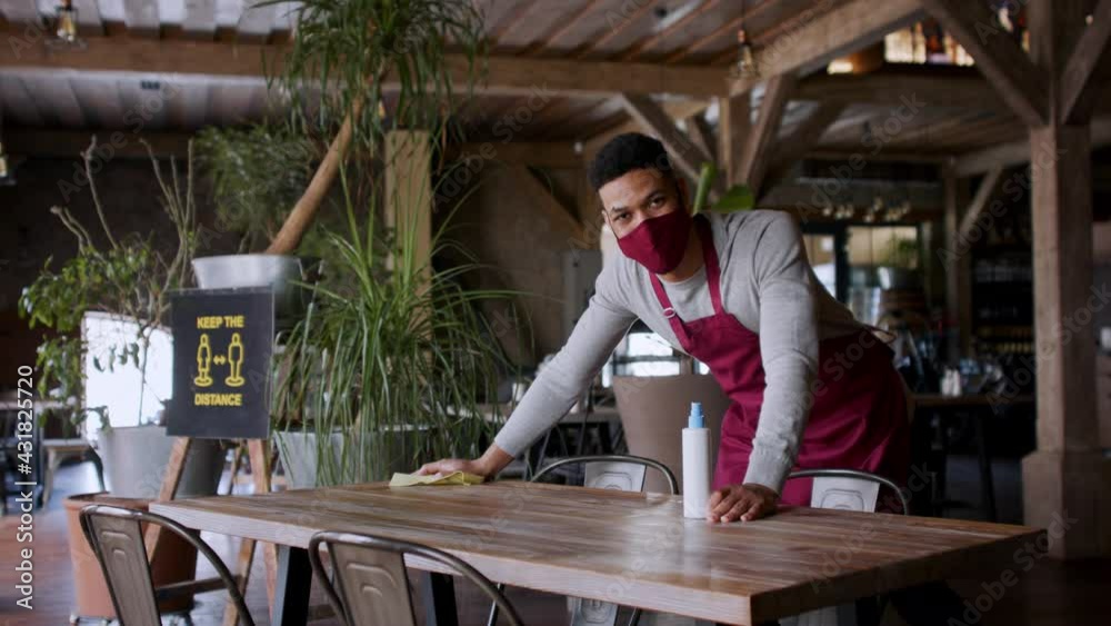 Waiter disinfecting tables in restaurant, looking at camera. Coronavirus concept. Waiter disinfecting tables in restaurant, looking at camera. Coronavirus concept.