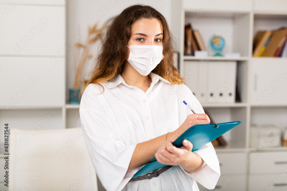Confident business woman in white protective mask holding folder in modern office