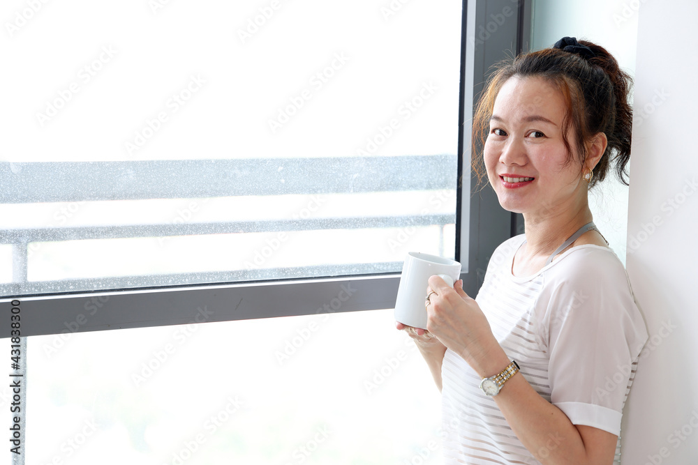 Beautiful attractive middle age southeast Asian woman standing next to daylight window curtain drink coffee from cup look at camera Beautiful attractive middle age southeast Asian woman standing next to daylight window curtain drink coffee from cup look at camera
