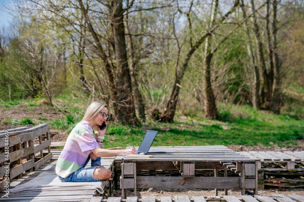 Fototapeta premium Blonde woman works outdoors using laptop and cell phone in beautiful sunny weather.