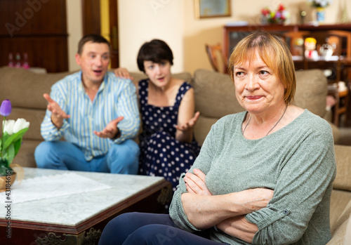 Portrait of upset offended elderly woman sitting on sofa at home on background with couple berating her.