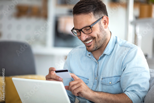 Young man shopping online with credit card at home. Mature casual man using laptop while looking at invoice. Smiling latin man managing finance with bills and laptop while sitting on couch at home.