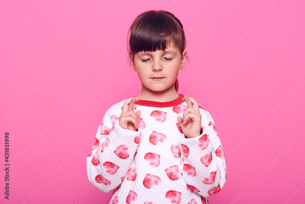Charming concentrated female kid wearing casual jumper with hearts keeping eyes closed and fingers crossed, hopes for better, wish dream comes true, isolated over pink background. Charming concentrated female kid wearing casual jumper with hearts keeping eyes closed and fingers crossed, hopes for better, wish dream comes true, isolated over pink background.