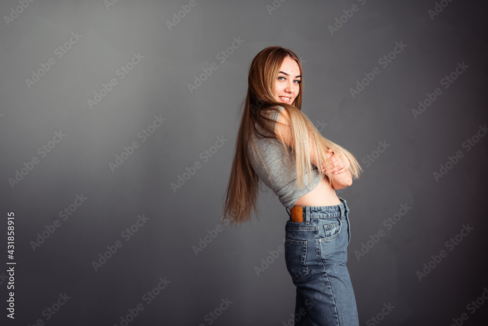 Long-haired blonde in jeans against the background of a gray wall hugs herself and smiles sincerely Long-haired blonde in jeans against the background of a gray wall hugs herself and smiles sincerely