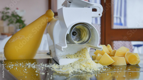 close-up of grated parmesan cheese coming down from the rotating electric grater with the background of an appetizing cheese wheel and on the sides the pieces ready to be grated photo