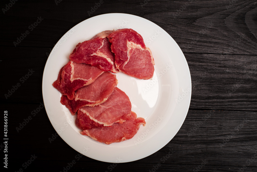 Raw pork chop on a white plate on a black wooden background. Fatty meat beaten off for cooking.